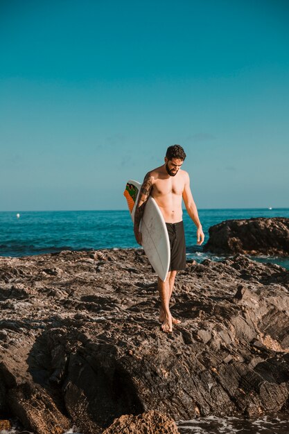 Jonge man met surfplank gaan op stenen wal in de buurt van water
