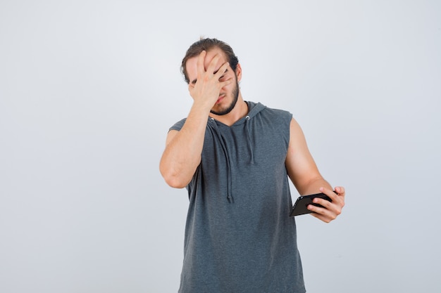 Jonge man met telefoon in de hand, gezicht bedekt met hand in t-shirt met een kap en op zoek ongelukkig, vooraanzicht.