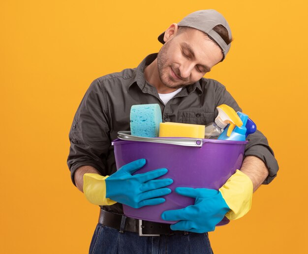 Jonge man met vrijetijdskleding en pet in rubberen handschoenen met emmer met reinigingsgereedschap kijken naar tools met liefde blij en tevreden staande over oranje muur