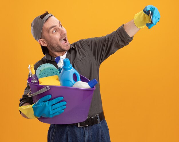 Jonge man met vrijetijdskleding en pet in rubberen handschoenen met emmer met reinigingsgereedschap met smartphone selfie doen blij en opgewonden staande over oranje muur