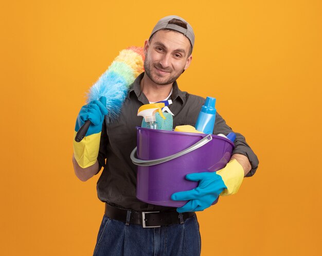 Jonge man met vrijetijdskleding en pet in rubberen handschoenen met emmer met schoonmaakgereedschap en kleurrijke stofdoek op zoek glimlachend klaar om schoon te maken staande over oranje muur