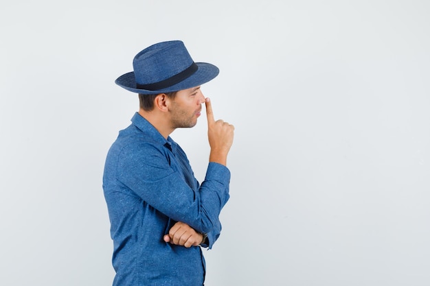 Jonge man neus aanraken met vinger in blauw shirt, hoed en peinzend kijken.