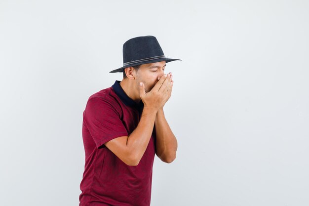 Jonge man niest en heeft griep in t-shirt, hoed en ziet er ziek uit. vooraanzicht.