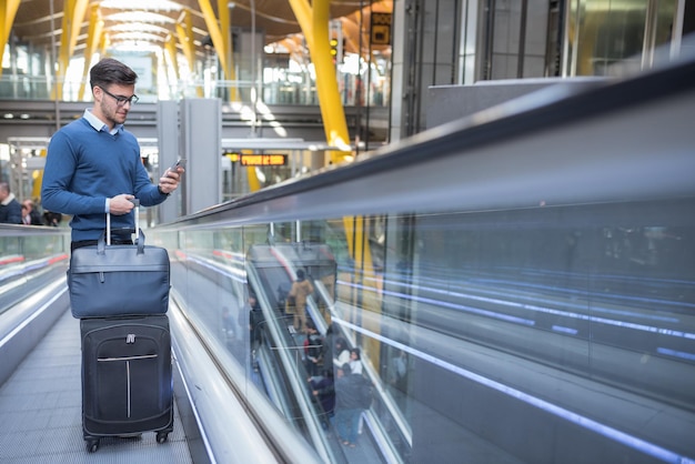 Jonge man op de roltrap op de luchthaven met behulp van zijn mobiele telefoon met zijn bagage glimlachen