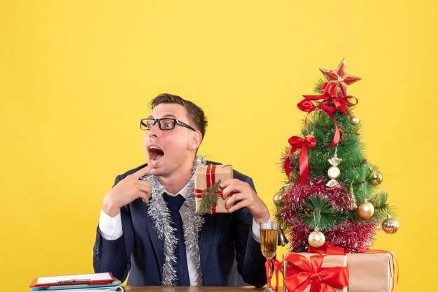 jonge man opening mond houden geschenk zitten aan de tafel in de buurt van kerstboom en presenteert op geel