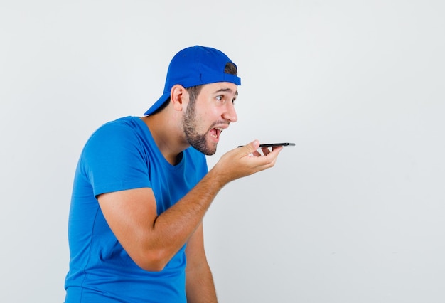 Jonge man opnemen van spraakbericht op telefoon in blauw t-shirt en pet vooraanzicht.