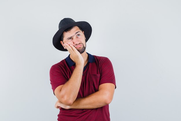 Jonge man opzij kijken in rood t-shirt, hoed en peinzend kijken. vooraanzicht.