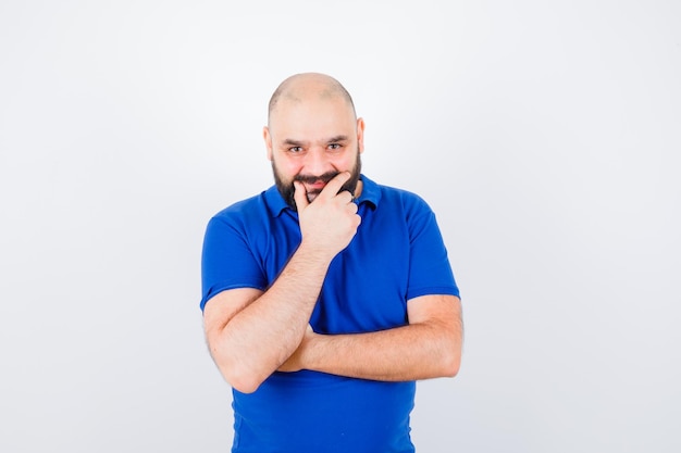 Jonge man poseren met de hand op zijn lippen, glimlachend in blauw shirt vooraanzicht.