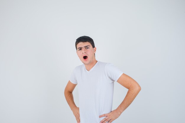 Jonge man poseren met de handen op de taille tijdens het openen van de mond in een t-shirt en op zoek verbaasd, vooraanzicht.