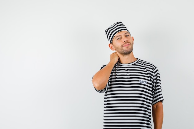 Jonge man poseren met hand op nek in gestreept t-shirt, hoed en elegant op zoek.
