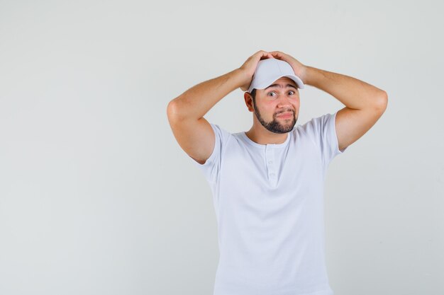 Jonge man poseren terwijl hij hand op zijn hoofd in wit t-shirt, pet en op zoek zelfverzekerd