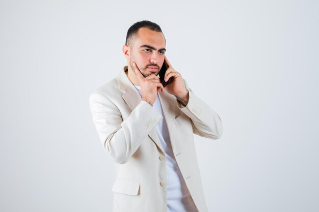 Jonge man praat met de telefoon en leunt wang bij de hand in wit t-shirt, jas en kijkt gefocust, vooraanzicht.