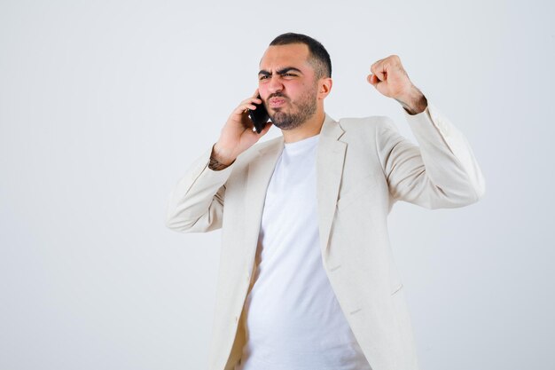Jonge man praat met iemand via de telefoon, balt zijn vuist in wit t-shirt, jas en kijkt woedend, vooraanzicht.