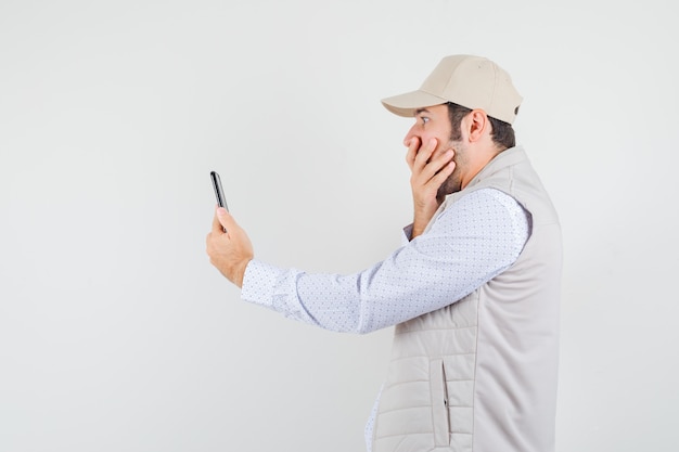 Jonge man praat met iemand via videocall en bedekt mond met hand in beige jas en pet en kijkt verbaasd. vooraanzicht.