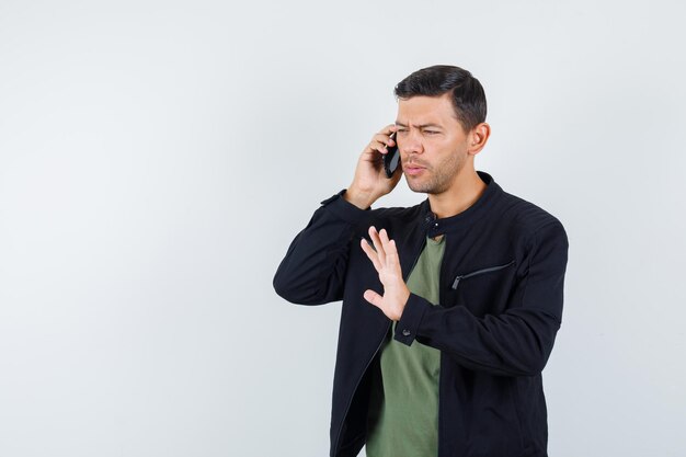 Jonge man praten op mobiele telefoon met stopgebaar in t-shirt, jas vooraanzicht.