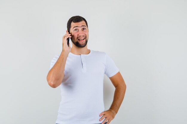 Jonge man praten over de telefoon in wit t-shirt en op zoek spraakzaam, vooraanzicht.