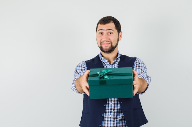 Jonge man presenteert geschenkdoos in overhemd, vest en kijkt zacht.