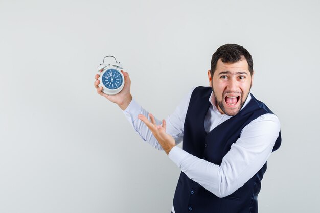 Jonge man schreeuwen terwijl hij op wekker in shirt en vest wijst en gehaast kijkt