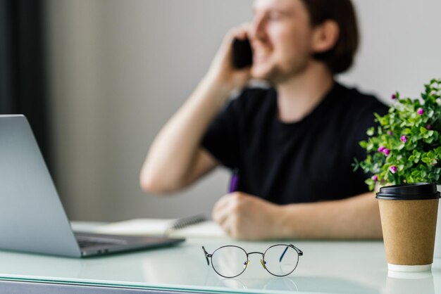 Jonge man schrijft notities terwijl hij telefoneert en thuis een laptop gebruikt