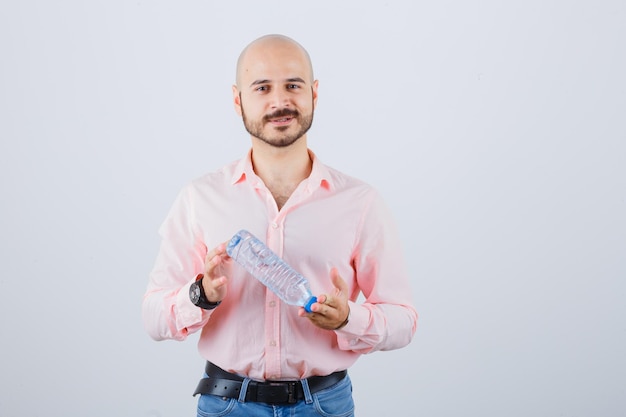 Jonge man schudden waterfles in roze shirt, jeans vooraanzicht.