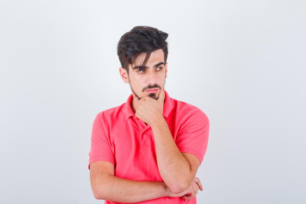 Jonge man staat in denkende pose in t-shirt en ziet er verstandig uit, vooraanzicht.