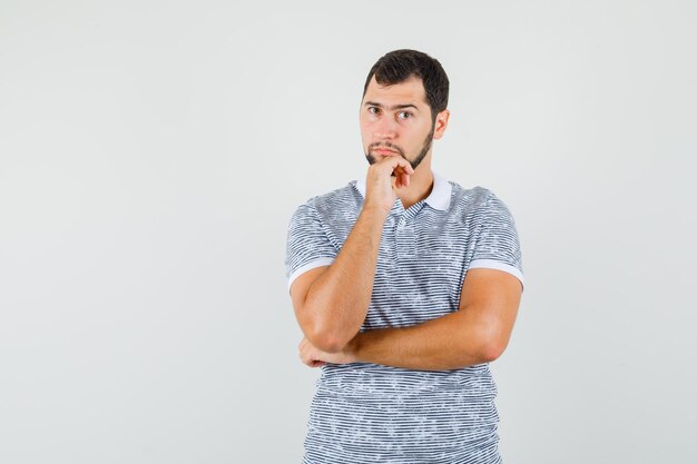 Jonge man staat in denkende pose in t-shirt en ziet er verstandig uit, vooraanzicht.