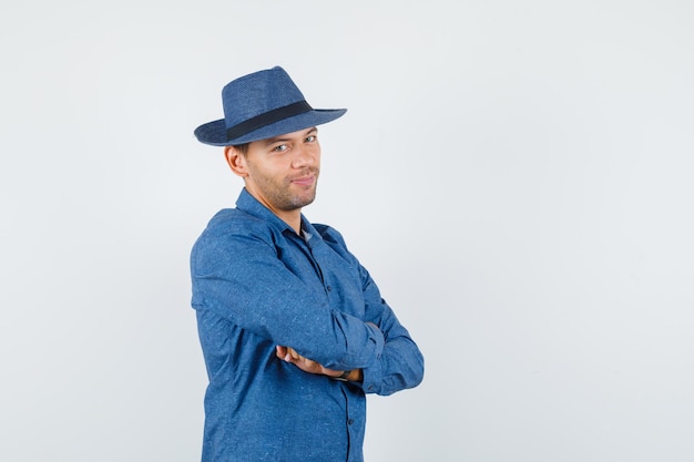 Jonge man staat met gekruiste armen in blauw shirt, hoed en ziet er vrolijk uit. vooraanzicht.