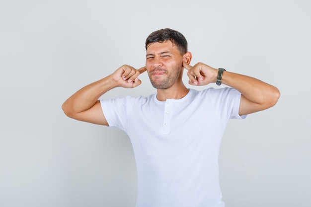 Jonge man steekt plug vingers in de oren in wit t-shirt en kijkt stressvol, vooraanzicht.