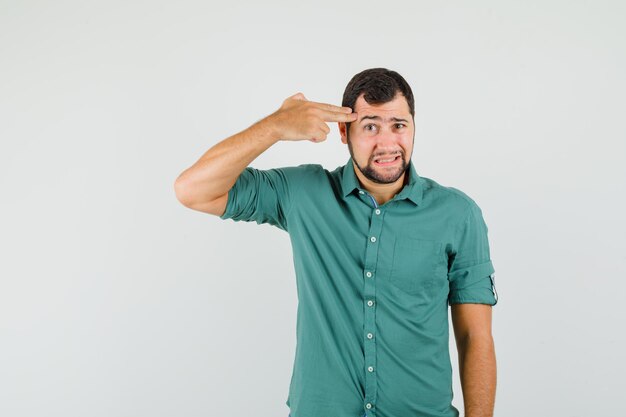 Jonge man toont pistoolgebaar naar zijn hoofd in groen shirt en kijkt verveeld. vooraanzicht.