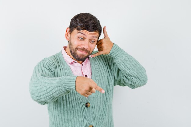 Jonge man toont telefoongebaar, wijst naar beneden in shirt, vest en kijkt gefocust, vooraanzicht.