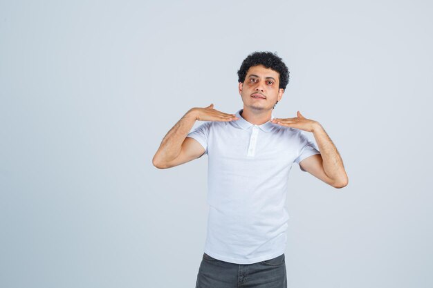 Jonge man toont zichzelf met handen in wit t-shirt, broek en ziet er trots uit, vooraanzicht.