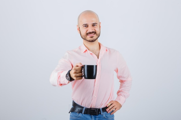 Jonge man toont zijn kopje thee in roze shirt, spijkerbroek en ziet er tevreden uit. vooraanzicht.