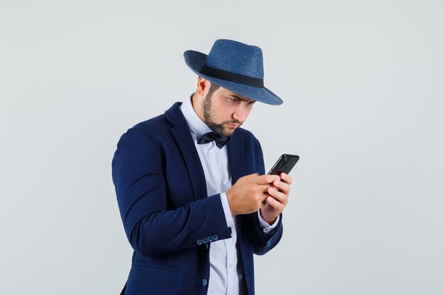 Jonge man typen op mobiele telefoon in pak, hoed en bezig op zoek. vooraanzicht.