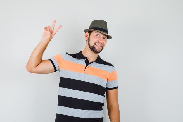 Jonge man v-teken in t-shirt, hoed tonen en vrolijk kijken.