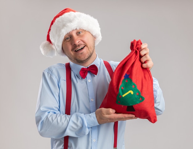 Gratis foto jonge man, vervelend, bretels, vlinderdas, in, kerstmuts, weergeven, rode kerstman, zak, vol, van, geschenken, kijken naar van fototoestel, vrolijke, en vrolijk, het glimlachen, staand, op, witte achtergrond