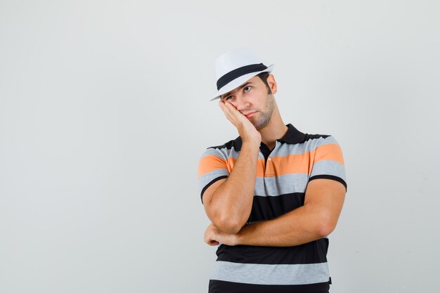 Jonge man wegkijken terwijl leunend op zijn handpalm in t-shirt, hoed en op zoek naar vermoeide ruimte voor tekst