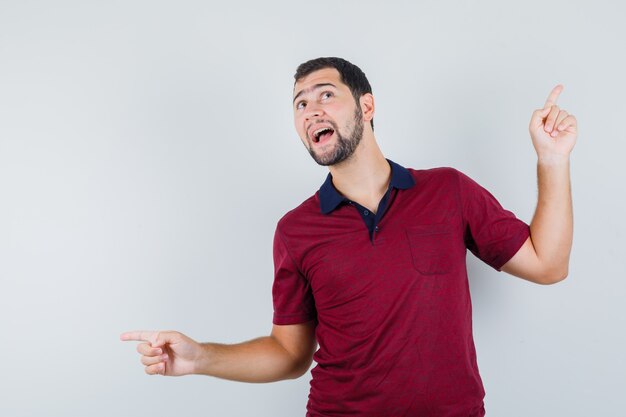 Jonge man wijst aan beide kanten in rood t-shirt en kijkt vrolijk, vooraanzicht.