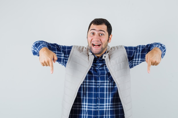 Jonge man wijst naar beneden in shirt, mouwloos jasje en kijkt blij, vooraanzicht.