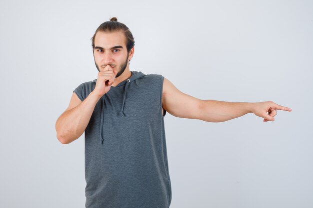 Jonge man wijst naar de zijkant, houdt hand op mond in t-shirt met een kap en ziet er knap uit, vooraanzicht.