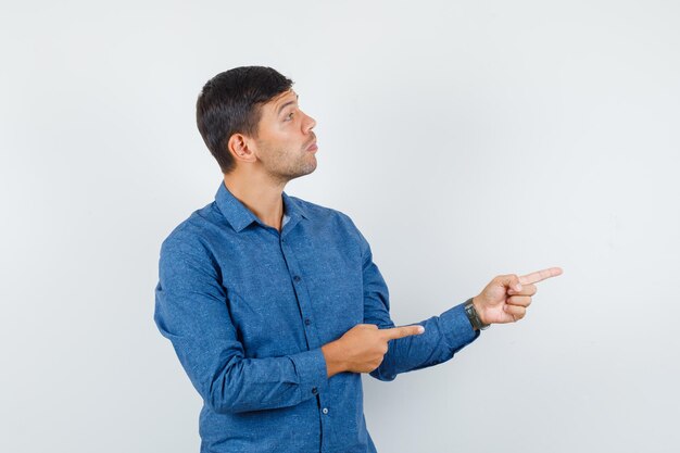 Jonge man wijst naar de zijkant in blauw shirt en kijkt gefocust. vooraanzicht.