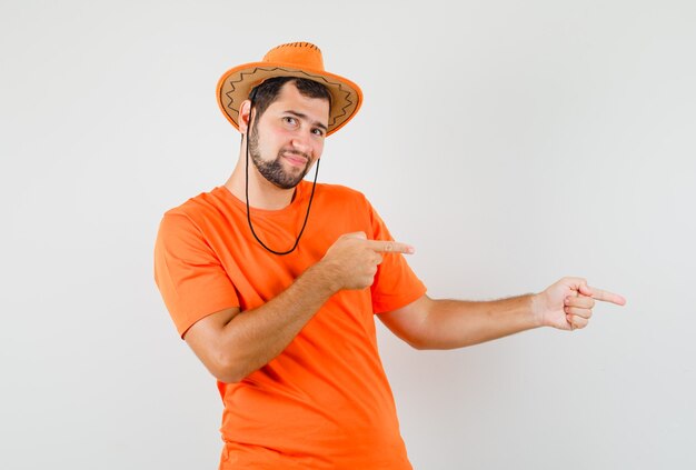 Jonge man wijst naar de zijkant in oranje t-shirt, hoed en ziet er vrolijk uit. vooraanzicht.