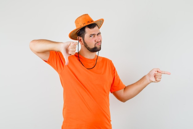 Jonge man wijst naar de zijkant, toont duim in oranje t-shirt, hoed en kijkt ontevreden. vooraanzicht.