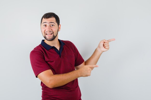 Jonge man wijst naar kant in rood t-shirt en kijkt blij. vooraanzicht.