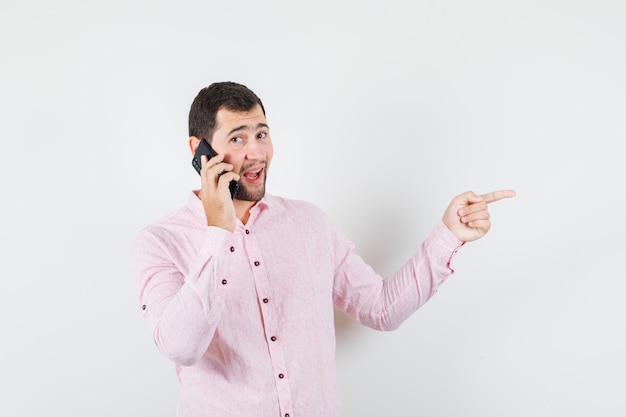 Jonge man wijst naar kant tijdens het praten op mobiele telefoon in roze shirt vooraanzicht.