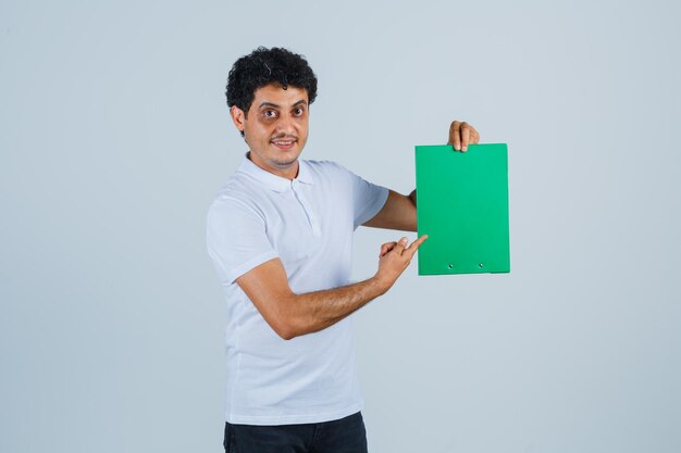 Jonge man wijst naar klembord in wit t-shirt, broek en ziet er vrolijk uit, vooraanzicht.