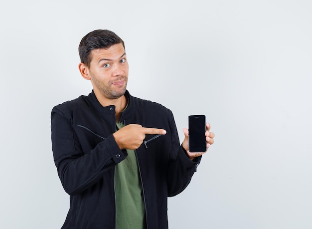 Jonge man wijst naar mobiele telefoon in t-shirt, jas en ziet er vrolijk uit. vooraanzicht.