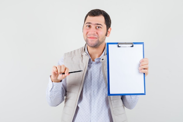 Jonge man wijst naar notebook met pen in beige jas en kijkt optimistisch, vooraanzicht.