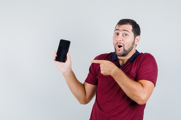 Jonge man wijst naar telefoon in rood t-shirt en kijkt verbaasd, vooraanzicht.