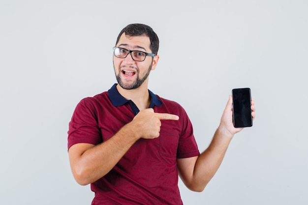 Jonge man wijst naar telefoon in rood t-shirt, optische bril en kijkt tevreden. vooraanzicht.