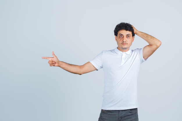 Jonge man wijst opzij, hand op het hoofd in wit t-shirt, broek en peinzend kijkend. vooraanzicht.